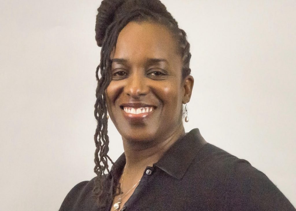 Headshot of Jovanka Beckles, CA Senate D7 Candidate; smiling woman in dark shirt, open at the collar to show a delicate silver and pearl pendant, dark hair in topknot with tendrils hanging down one side