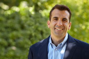 Head and shoulders shot of smiling Sam Liccardo in dark blue suit jacket with lighter blue shirt, no tie, in front of green shrubbery