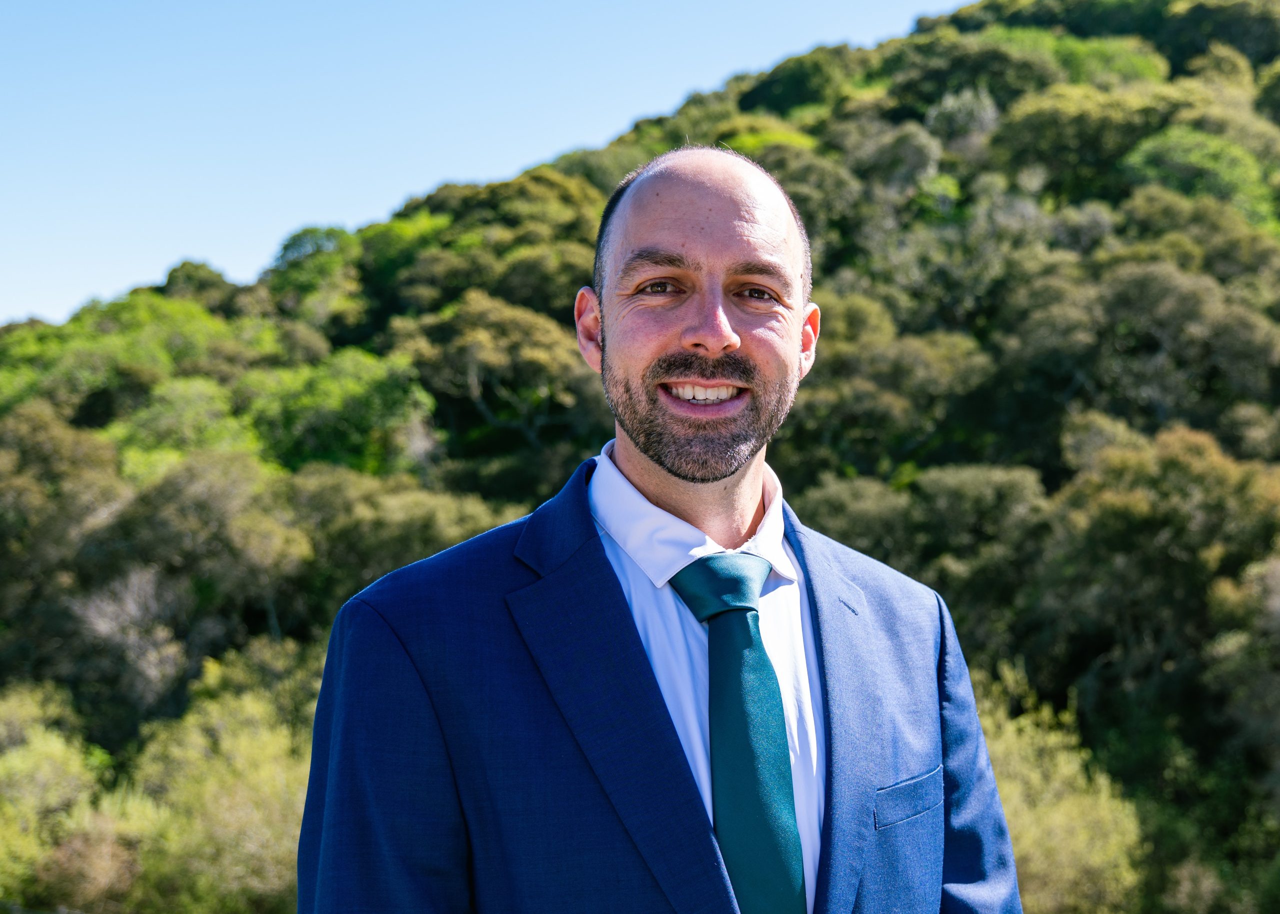 Adam Loraine for San Mateo City Council District 5. Adam in a blue suit and tie, a green hillside behind hijm. Photo credit: Christopher Joseph Ball, used with permission