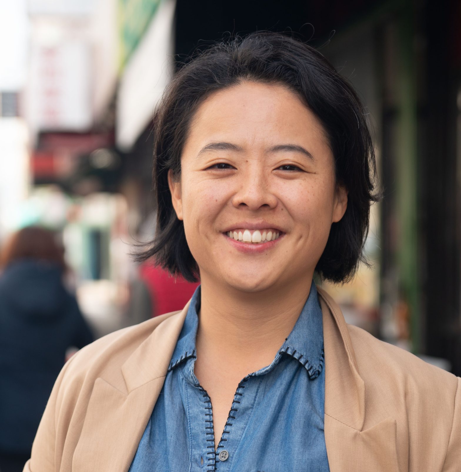 Charlene Wang for Oakland City Council district 2. Charlene is smiling at the camera, a blurred background of China Town, Oakland. Charlene Wang, used with permission.