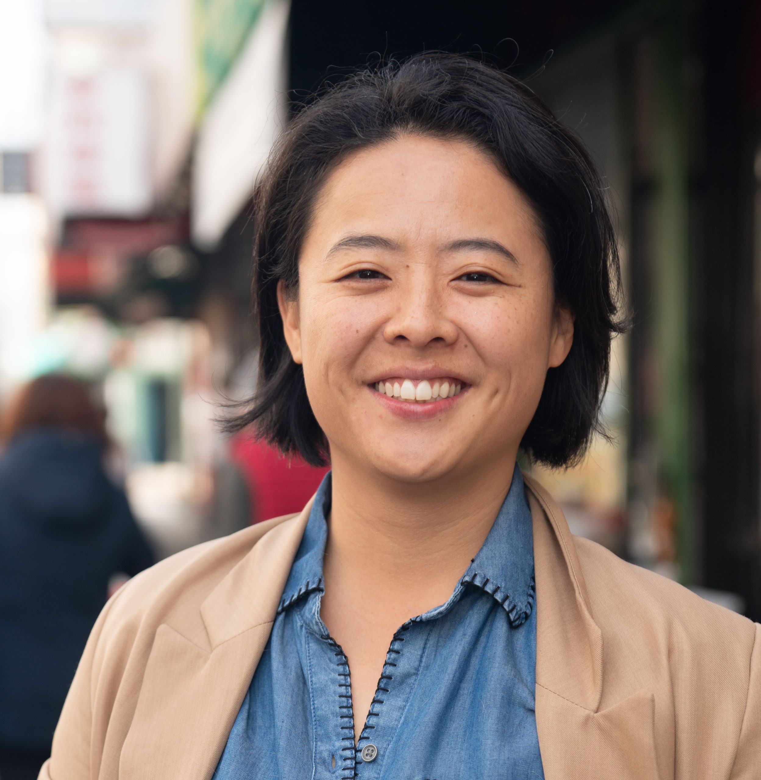 Charlene Wang for Oakland City Council district 2. Charlene is smiling at the camera, a blurred background of China Town, Oakland. Charlene Wang, used with permission.