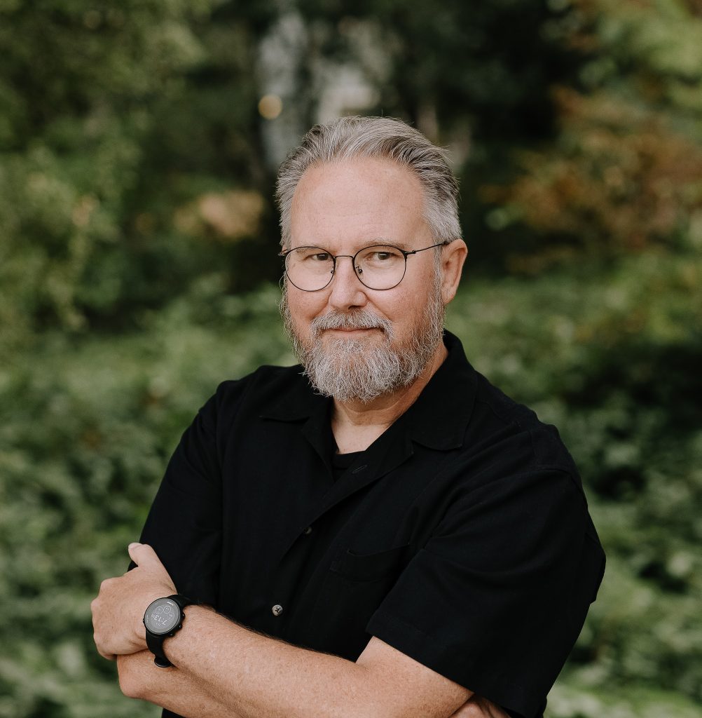 Chris Herrod for Healdsburg district 4. Man in black shirt sitting, arms folder, warm glint in his eyes. Photo credit: Chris Herrod, used with permission