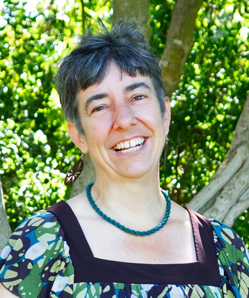 Daria Wrubel for Berkeley City Council district 1. Woman in a colorful patterned blouse and a blue necklace with a big smile, and with a tree behind her. Photo credit: Daria Wrubel, used with permission.