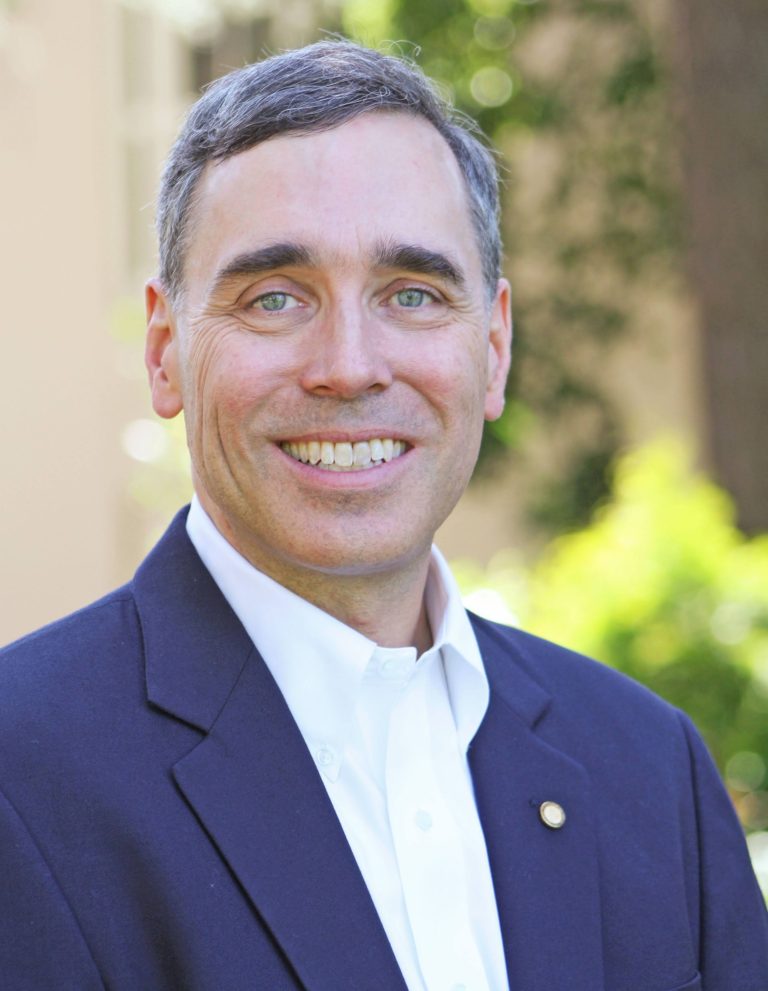 Paul Donahue for Mountain View City Council. Man in a navy blue jacket with blue-green eyes is smiling at the camera, trees behind him. Photo credit: Paul Donahue, used with permission.