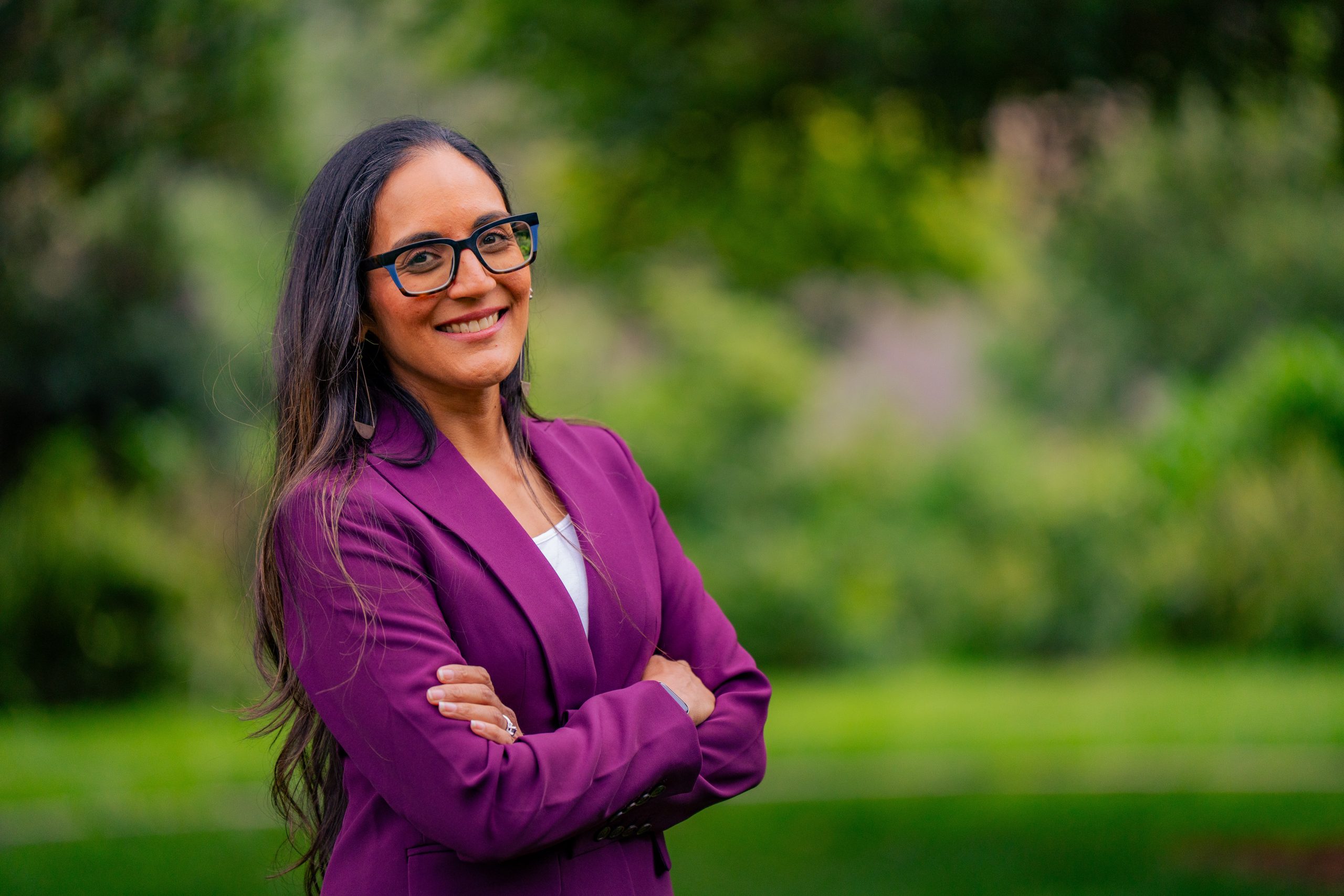 Seema Patel for San Mateo City Council District 3. Woman with long hair and wearing a fuchsia colored jacket, smiling, a green park behind her. Photo credit: Seema Patel, used with permission.