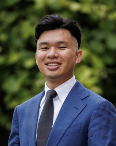 Cameron Sasai for Contra Costa Community College District, Ward 1 Man in a blue suit and gray tie, smiling, and standing in front of a tree. Photo credit: Cameron Sasai, used with permission.