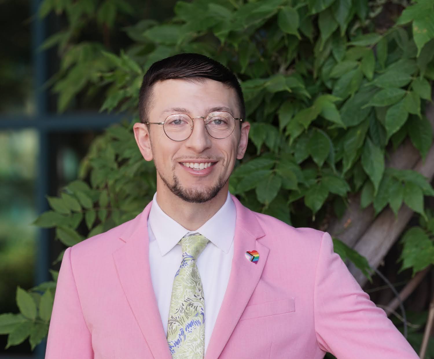 Chris Sturken for Redwood City District 2. Man in a pink suit jacket and pale tie with a rainbow pin, stands smiling in front of a tree. Photo credit: Chris Sturken, used with permission.