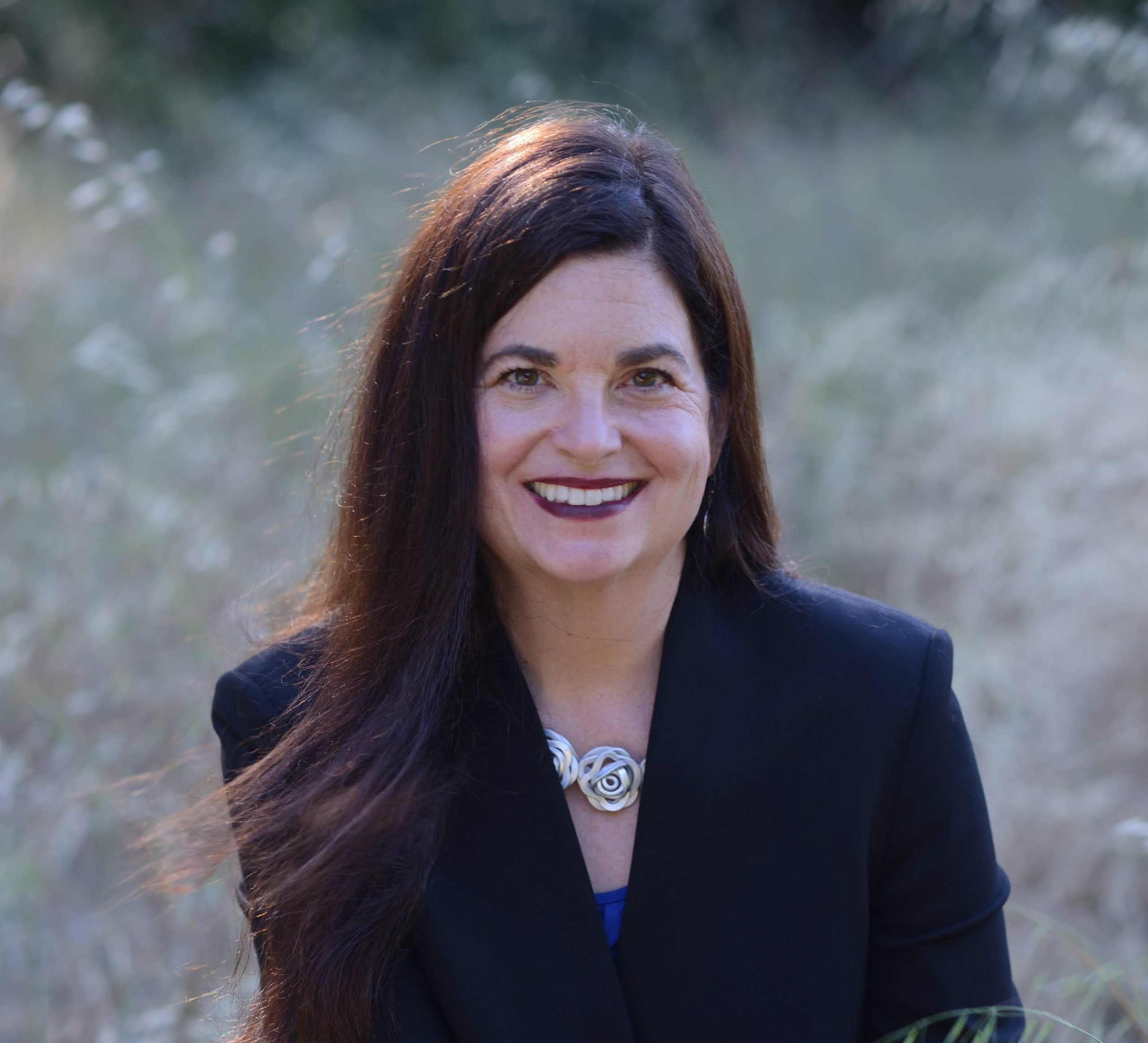 IdaRose Sylvester for Mountain View City Council. A woman, wearing a blue jacket and a silver necklace of "roses", is standing in a field with her long hair blowing in wind. Photo credit: IdaRose Sylvester, used with permission.