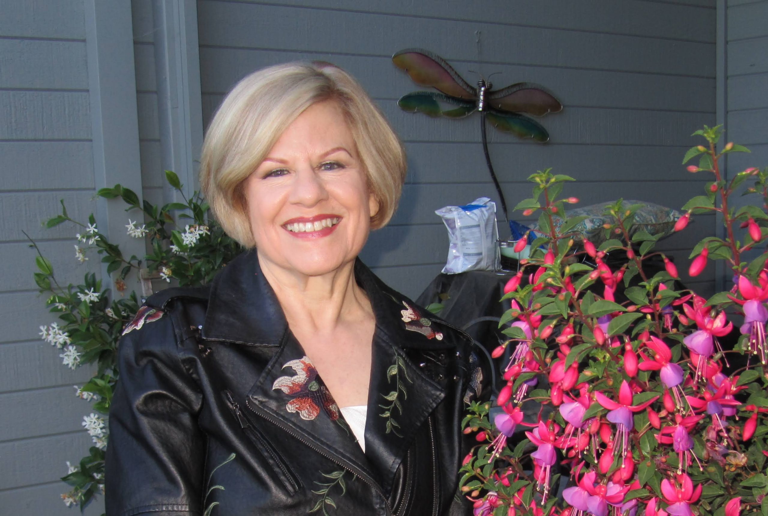 Joan Cox for Sausalito City Council. Woman with black jacket with flowers on it standing on a porch with flowering plants. She is smiling. Photo credit: Joan Cox, used with permission.